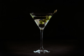 Glass of classic dry martini cocktail with olives on dark stone table against a black background.