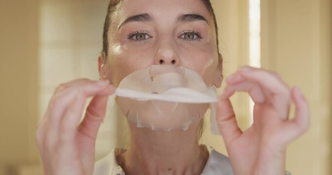 Beauty shot of mature woman is taking off paper sheet mask from her purified and clean face touching it gently and smiling in camera on golden background. Concept of skincare, cosmetics, healthcare