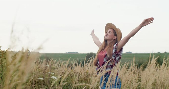 Satisfied Adult Girl standing on the Field, Smiling and Rises Hands up. Happy Woman enjoying of Nature, looking at the Sky, wearing Straw Hat and Denim Skirt. Having Positive Emotions.