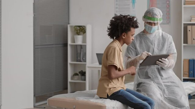 Medium long of doctor wearing protection suit, gloves, mask, face shield and African school-aged male patient sitting on medical couch in office, using tablet computer for filling in questionnaire