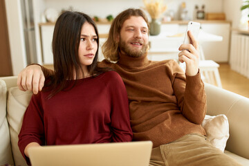 Young unshaven male holding mobile phone, scrolling newsfeed on social network, sitting comfortably next to his annoyed girlfriend who is trying to work using laptop. People, relationships and gadgets