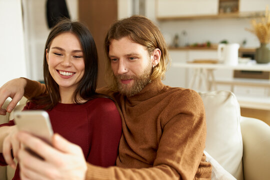 Fashionable young Caucasian man with beard enjoying free time indoors, having fun with his cute wife, laughing, taking selfie on mobile phone, using face effects and filters on social network