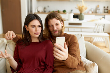 Human emotions, technology and relationships concept. Portrait of frowning young female making emotional gesture being angry while boyfriend showing her annoying video or meme on smart phone
