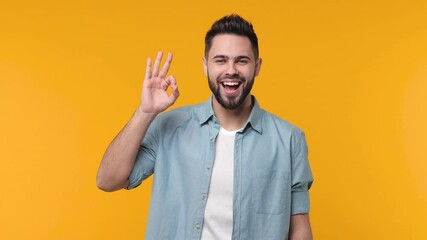 Cheerful laughing bearded young man 20s years old in basic blue shirt white t-shirt isolated on yellow background in studio. People sincere emotions lifestyle concept. Showing ok okay gesture blinking
