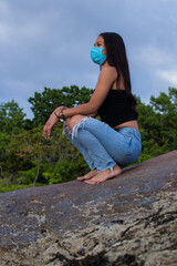 Vertical shot of an attractive Hispanic female with long hair wearing a face mask squatting on rocks