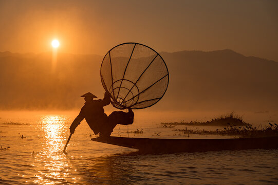 INTHA FISHERMEN, INLE LAKE, SHAN STATE, MYANMAR - 18 January 2020: Traditional fishing technique balancing on one leg on a narrow boat using a conical net at sunrise.