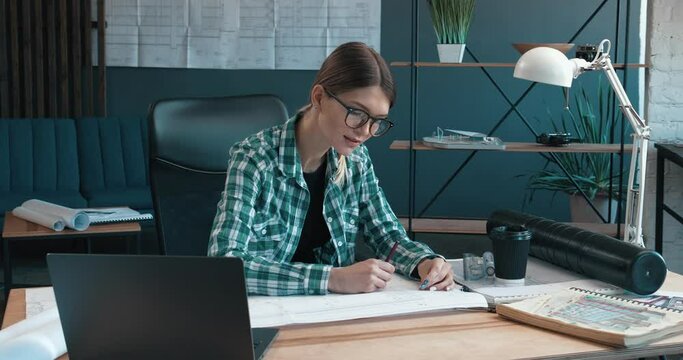 Attractive Woman Architect Making drawings with pencil in Modern Office. Looking at the camera with a Charming Smile in trendy eyeglasses and dressed in Shirt. Portrait.