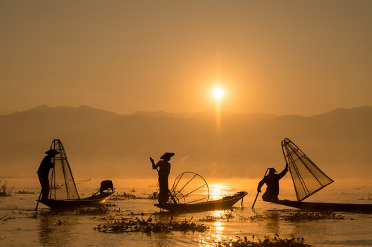 INTHA FISHERMEN, INLE LAKE, SHAN STATE, MYANMAR - 18 January 2020: Three traditional fishermen using traditional conical net technique silhouetted at sunrise.