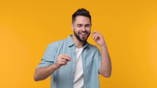 Cheerful funny bearded young man 20s years old in basic blue shirt isolated on yellow color background in studio. People sincere emotions lifestyle concept. Dancing clenching fists waving rising hands