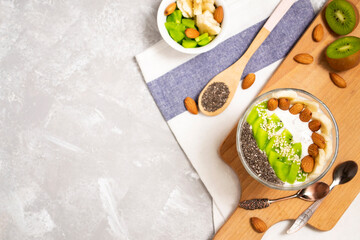 There is a bowl of chia pudding with kiwi, banana and assorted seeds on a wooden board. Healthy food concept on concrete background with copy space.