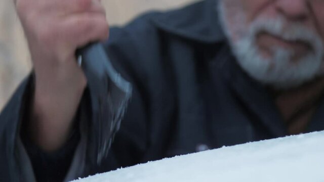 Senior person with grey beard hits automobile roof with black rubber ice scraper on cold winter day. Concept weather conditions