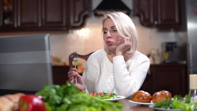 Bored carefree Caucasian woman chewing salad and watching movie on laptop as smelling unpleasant smell from kitchen. Portrait of stressed beautiful lady running to burnt food at background.