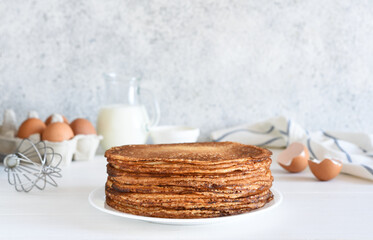 Fototapeta na wymiar A stack of thin pancakes and sour cream, a whisk, eggs and milk on a concrete background.
