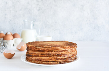 A stack of thin pancakes and sour cream, a whisk, eggs and milk on a concrete background.