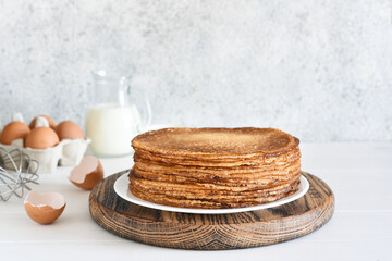 Fototapeta na wymiar A stack of thin pancakes and sour cream, a whisk, eggs and milk on a concrete background.