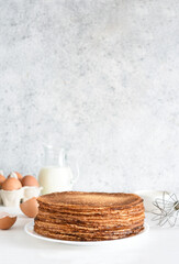A stack of thin pancakes and sour cream, a whisk, eggs and milk on a concrete background.