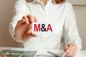 A woman in a white shirt holds a piece of paper with the text: M and A. M A - mergers and...