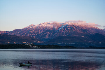 A fisherman is fishing in a boat in the Bay of Kotor, against the backdrop of the snow-covered Lovcen mountain at sunset.