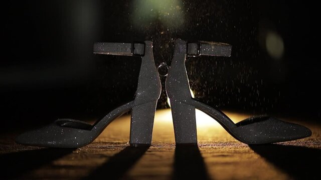 Wedding rings placed in between brides wedding shoes sprinkled with perfume spray on dark background. Slow motion. Pair of marriage symbols. Love of bride and groom, wife and husband. Matrimony symbol