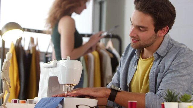 Interior of fashion studio with male clothes designer working at sewing machine talking with female colleague in background- shot in slow motion
