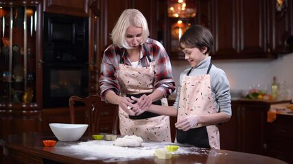 Mother stops son from eating raw dough. Portrait of smiling Caucasian blond woman kneading bakery as boy in apron helping parent in kitchen at home. Joyful family cooking and having fun.