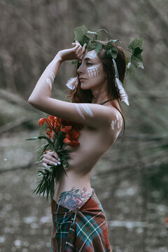 Mystical portrait of a mysterious Wiccan woman with tanned skin and long red hair posing in the woods. The young girl holds tiger lilies in her hands and has witchcraft patterns on her skin.