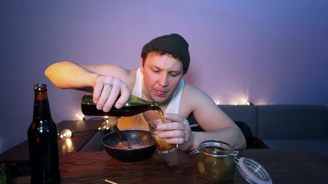 The man watches TV, drinks beer, eats sausages with cucumber and relaxes