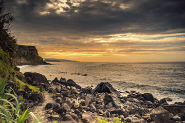Seascape with volcanic stones between the Atlantic Ocean and pedestrian path under dramatic sunset sky and cliffs on the horizon at Viola beach, São Miguel - Azores PORTUGAL