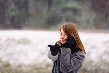 A cute girl of caucasian appearance is talking on the phone and looking at the camera on a snowy background.