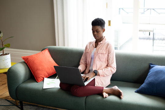 African american woman sitting on couch using laptop talking on smartphone working from home