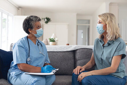 Senior african american female doctor talking with female patient both wearing face masks at home