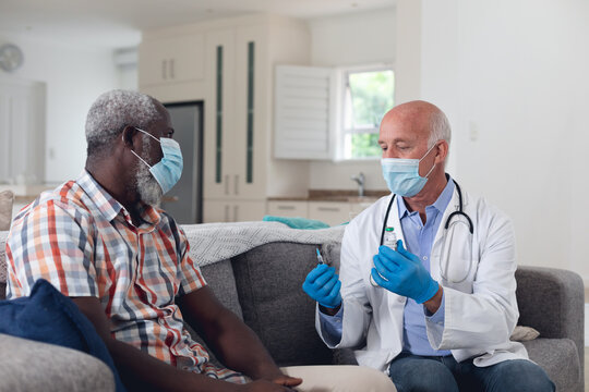Senior caucasian male doctor preparing vaccine for male patient both wearing face masks at home