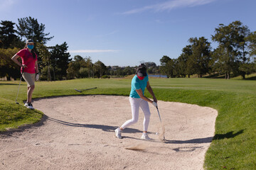 Two caucasian women wearing face masks playing golf one taking shot from a bunker