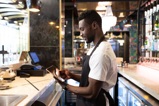 African american male barista standing behind a bar using a digital tablet and smiling