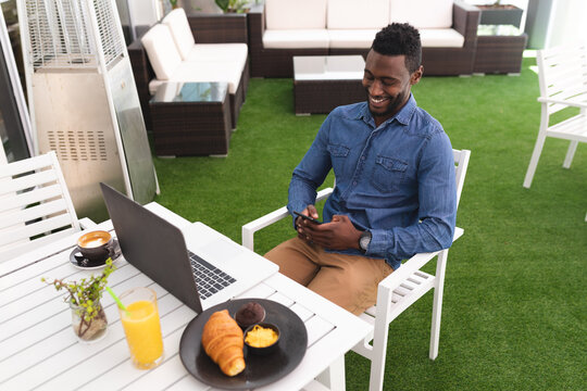 African american man sitting in a cafe using smartphone and laptop eating breakfast