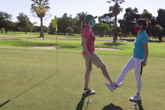Two caucasian women wearing face masks greeting on golf course by touching ankles