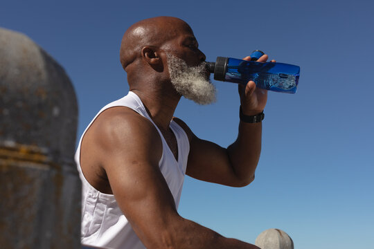 Fit senior african american man sitting drinking from water bottle against blue sky