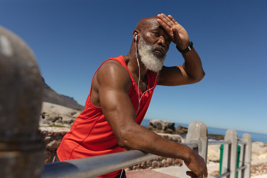 Fit senior african american man wearing earphones in sun by sea mopping brow