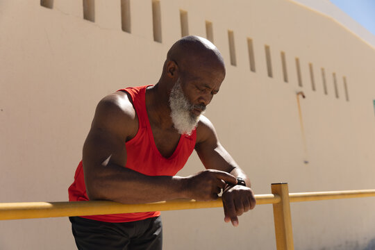 Fit senior african american man exercising using smartwatch leaning on fence in sun