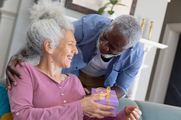 Senior mixed race couple in living room man giving woman a gift