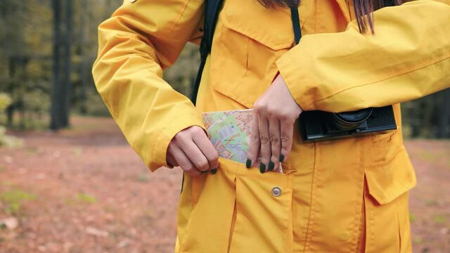 Happy girl folds map of area and puts it in pocket of yellow jacket and takes photo of nature on her camera.