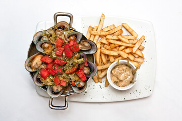 Mussels in a frying pan and on a plate with chips and sauce on white background, top view