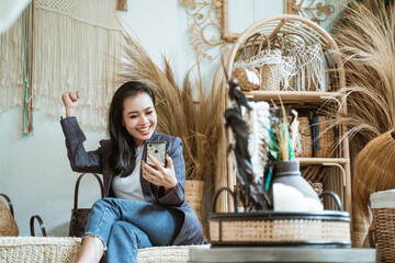 happiness woman in blazer with hand up while looks to screen smartphone in a handicraft shop on a...