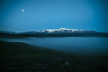 Atmospheric mountains landscape with dense fog and great snow mountain top under twilight sky. Alpine scenery with big snowy mountains over thick fog in night. High snow pinnacle above clouds in dusk.