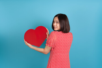 smiling beautiful young asian woman in red dress stands back and holds big red paper heart isolated on blue background.