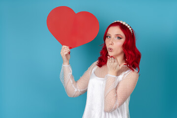 close-up a beautiful young woman in a white dress and with red hair holds a large red paper heart and blows a kiss isolated on a blue background.
