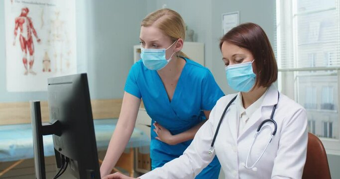 Caucasian medical professionals in protective msks working on computer in on-call room. Good-looking young nurse and adult female doctor making reports and looking at camera. Profession concept.