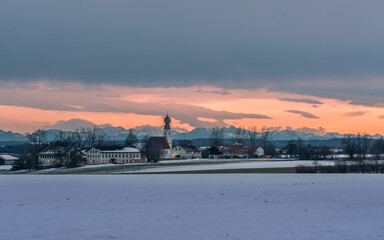 Kirch mit Gebäuden  vor Alpenpanorama bei Sonnenuntergang im Chiemgau im Winter mit Schnee und Bäumen