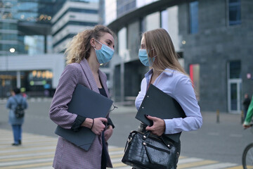 Two women in medical masks are talking on the street next to an office building