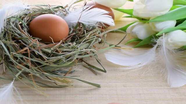 Color eggs in basket with spring tulips, white feathers on wooden table background in Happy Easter decoration. Foil minimalist egg design, modern top view design